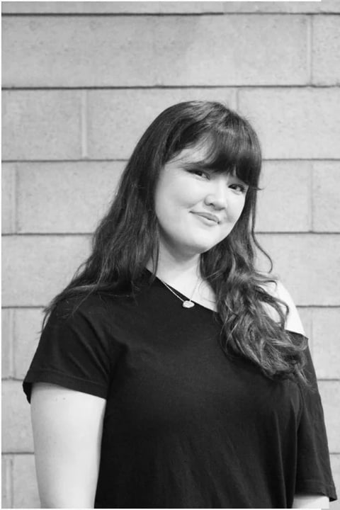 Black and white portrait of a smiling woman with long dark hair and bangs wearing a black t-shirt against a brick wall background