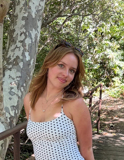 Young woman with brown wavy hair wearing a white polka dot top, posing outdoors by a birch tree with green foliage in background