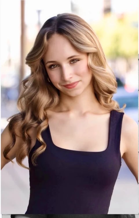 Woman with long wavy blonde hair wearing a black sleeveless top, smiling at camera in an outdoor urban setting