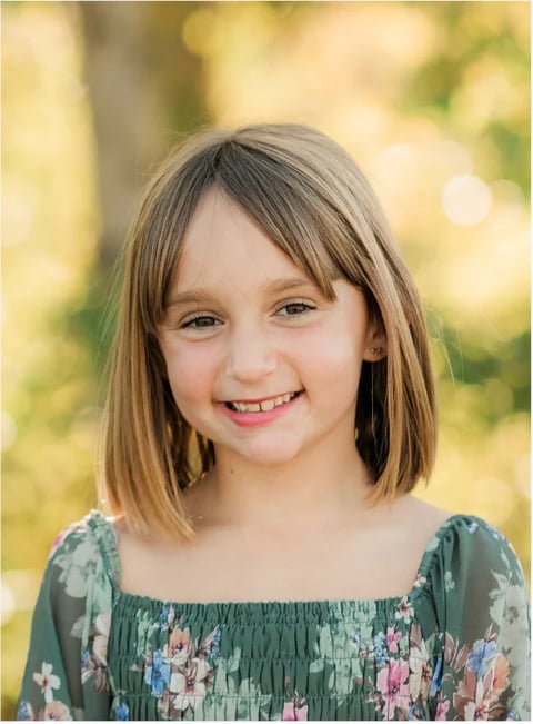 Young girl with shoulder-length brown hair smiling at camera, wearing a green floral dress against a blurred green background